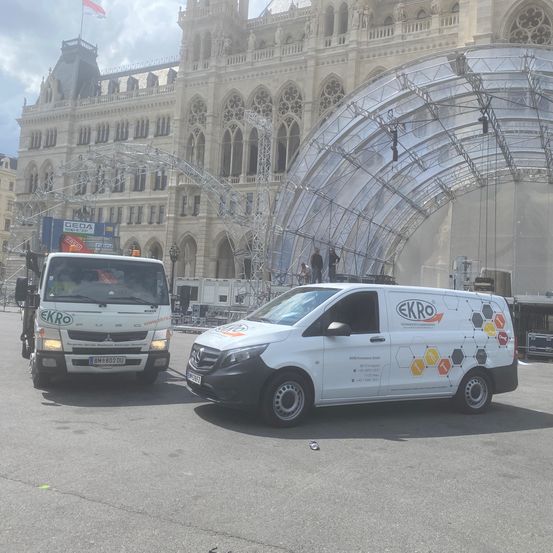 Zwei weiße Lieferwagen mit Firmenlogos vor einem großen, historischen Gebäude mit aufwendiger Architektur und einer temporären Struktur geparkt.