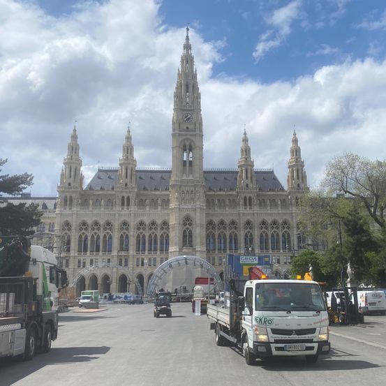 Ein LKW fährt an der Wiener Stadthalle vorbei, mit Statuen und Bäumen im Vordergrund.