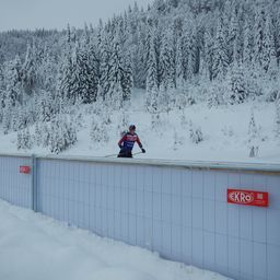 Ein Skifahrer in einem blauen und roten Outfit bereitet sich darauf vor, von einem verschneiten Berg vor einer Mauer mit einem roten ECRO-Schild zu springen.