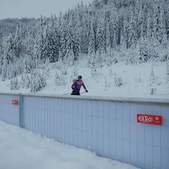 Ein Skifahrer in einem blauen und roten Outfit bereitet sich darauf vor, von einem verschneiten Berg vor einer Mauer mit einem roten ECRO-Schild zu springen.