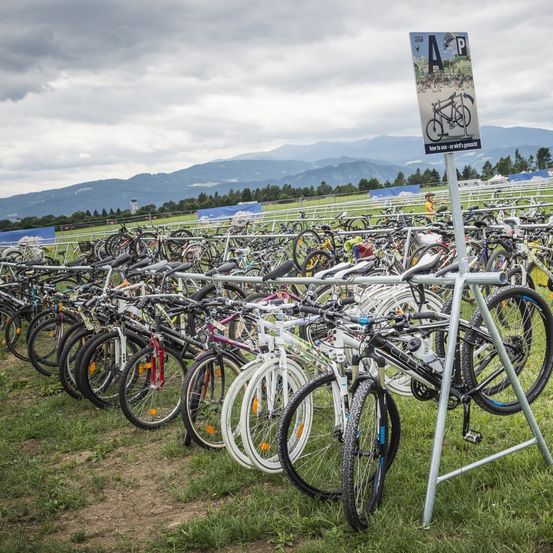 Eine Reihe von Fahrrädern ist auf einem Rasenfeld geparkt. Ein Schild mit der Aufschrift 'AP' und einem Bild eines Fahrrads steht auf einem Metallständer.
