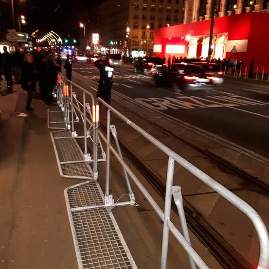 Ein Polizeibeamter steht neben einer Metallbarriere auf einer belebten Straße bei Nacht. Mehrere Menschen warten auf dem Bürgersteig, und Autos fahren vorbei. Der Hintergrund ist verschwommen mit Gebäuden, Straßenlaternen und Schildern.