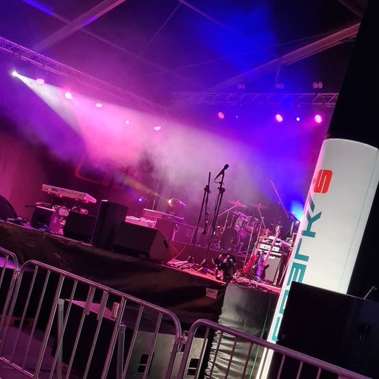 Eine Bühne mit mehreren Lichtern und Lautsprechern, mit einem großen weißen Banner, auf dem in großen orangefarbenen Buchstaben 'ARK' steht und darunter das Wort 'UN'.