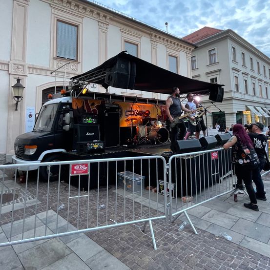 Eine Rockband tritt auf einer Bühne bei einer Stadtplatzveranstaltung auf. Ein schwarzer Lastwagen dient als Bühne mit einem großen Lautsprecher und einem Vordach. Ein Mann spielt Gitarre, ein anderer Schlagzeug und ein dritter singt. Zuschauer versammeln sich um sie herum und beobachten die Aufführung.
