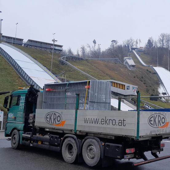 Ein Lkw mit dem Logo EKR0 steht neben einer verschneiten Skipiste mit Skisprungschanzen, mit einem grauen Auto in der Nähe.