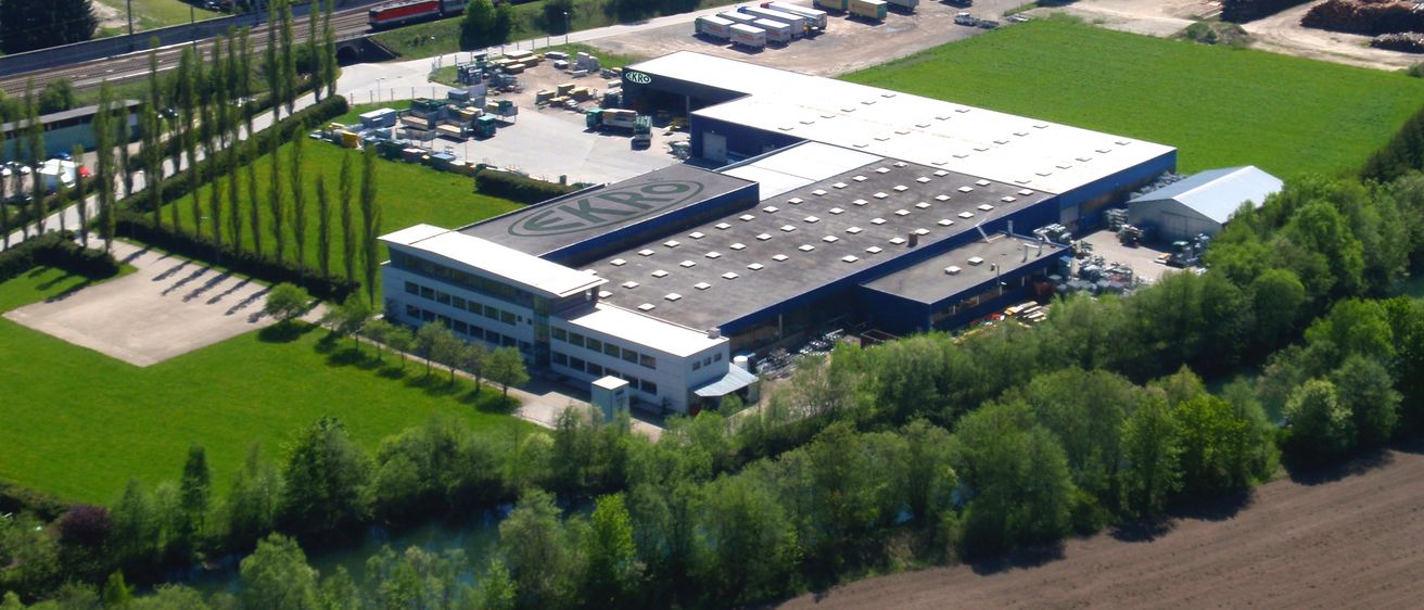 Luftaufnahme eines Industriegebiets mit einem großen Gebäude im Vordergrund und einem vorbeifahrenden Zug auf einer nahegelegenen Bahnstrecke.