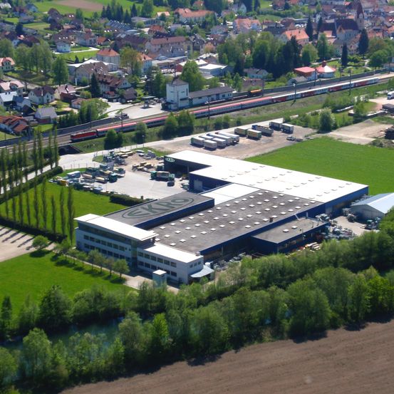 Luftaufnahme eines großen Industriegebäudes mit einem Logo auf dem Dach, umgeben von einem grünen Feld, mehreren Lastwagen und einem Zug auf den Gleisen.