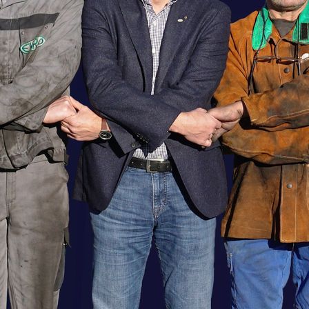 Drei Personen stehen eng beieinander, wobei die Frau auf der linken Seite einen roten Schal trägt und der Mann auf der rechten Seite eine Uhr trägt. Die mittlere Person ist in Arbeitskleidung mit einem grünen Logo auf der linken Brust gekleidet.