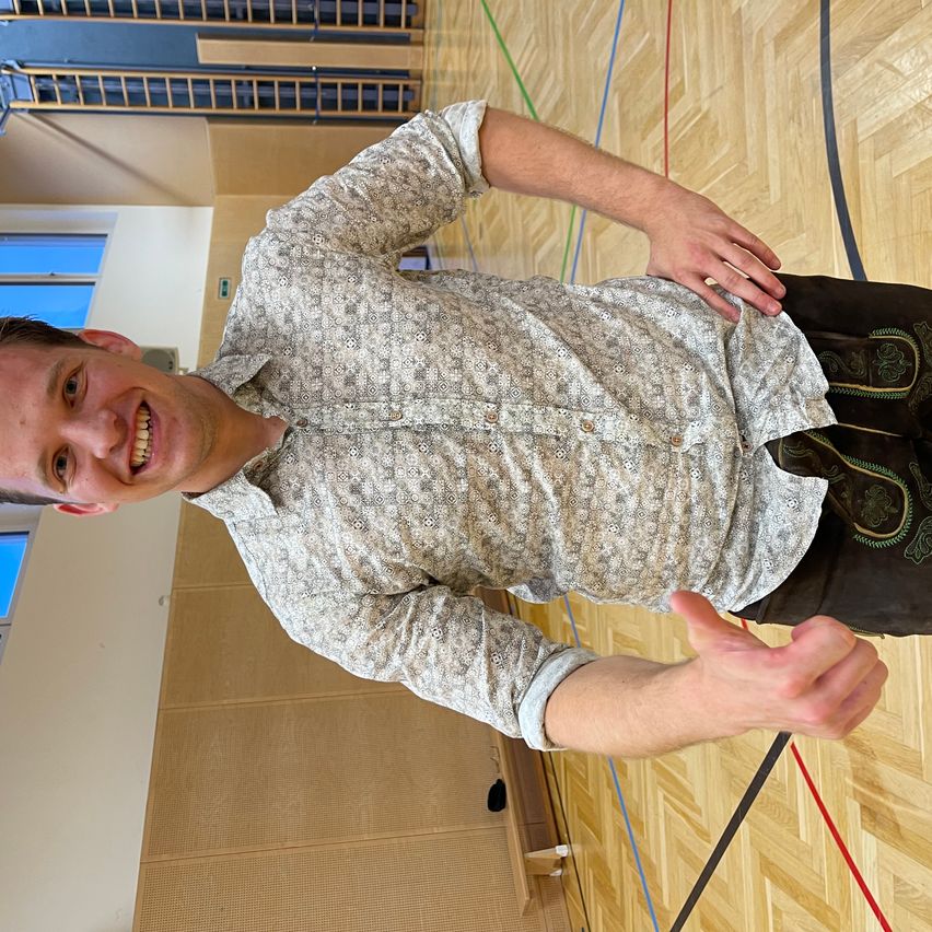 Ein Mann mit einem Lächeln auf dem Gesicht liegt auf einem Holzboden, wahrscheinlich in einer Turnhalle. Er trägt ein gemustertes Hemd und traditionelle Hosen.