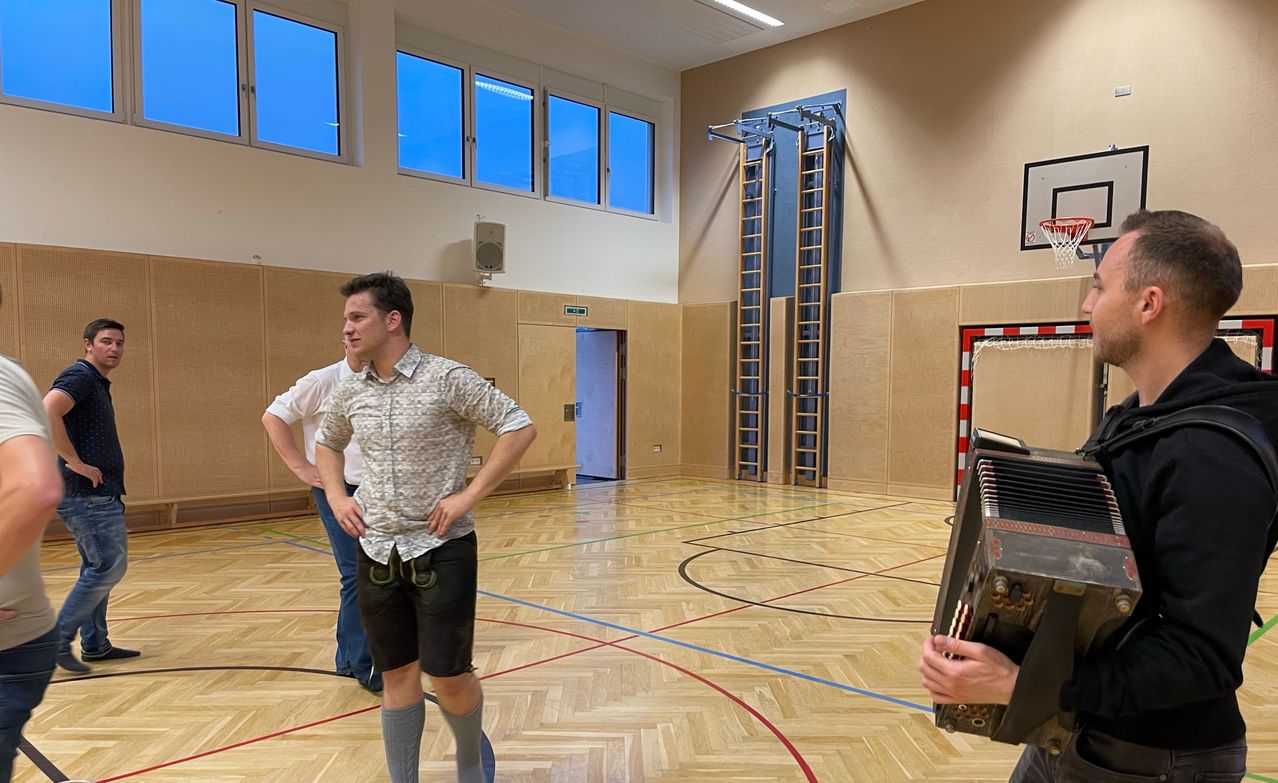 Zwei Männer in Shorts sind in einer Turnhalle, einer spielt Akkordeon und der andere beobachtet. Der Raum hat Holzböden, große Fenster und zwei an der Wand montierte Leitern.