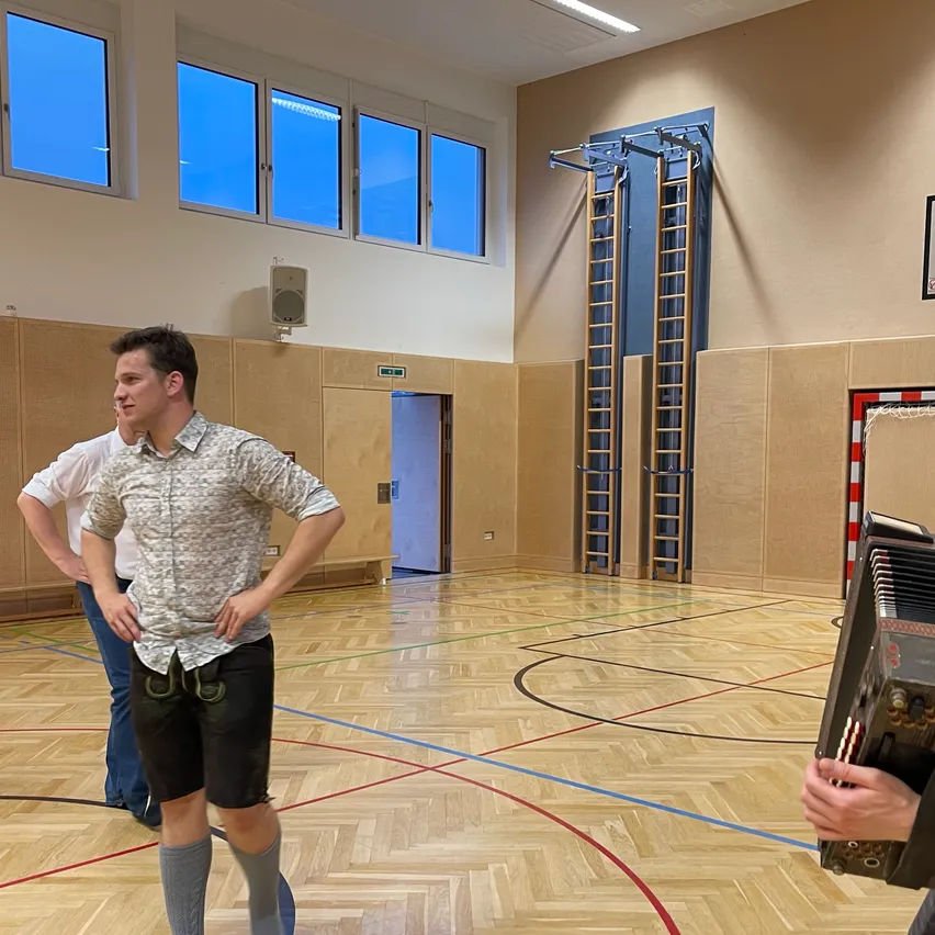 Zwei Männer in Shorts sind in einer Turnhalle, einer spielt Akkordeon und der andere beobachtet. Der Raum hat Holzböden, große Fenster und zwei an der Wand montierte Leitern.