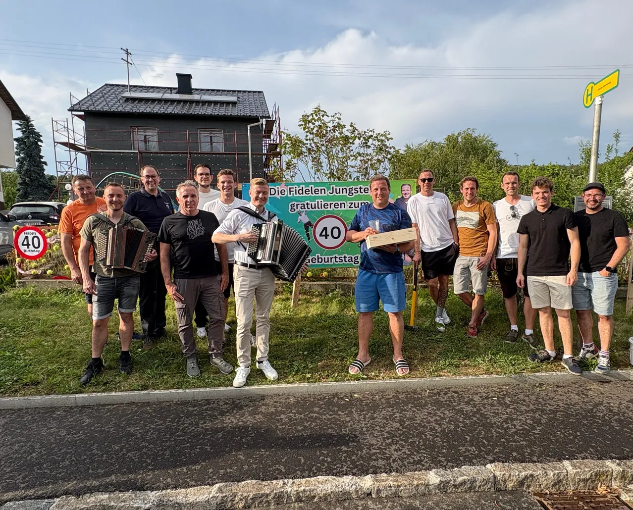 Eine Gruppe von Männern steht in der Nähe einer Straße, posiert für ein Foto mit einem Schild, das '40' und eine Geburtstagsbotschaft zeigt. Sie tragen Freizeitkleidung und einige halten Musikinstrumente. Hinter ihnen steht ein Haus im Bau.