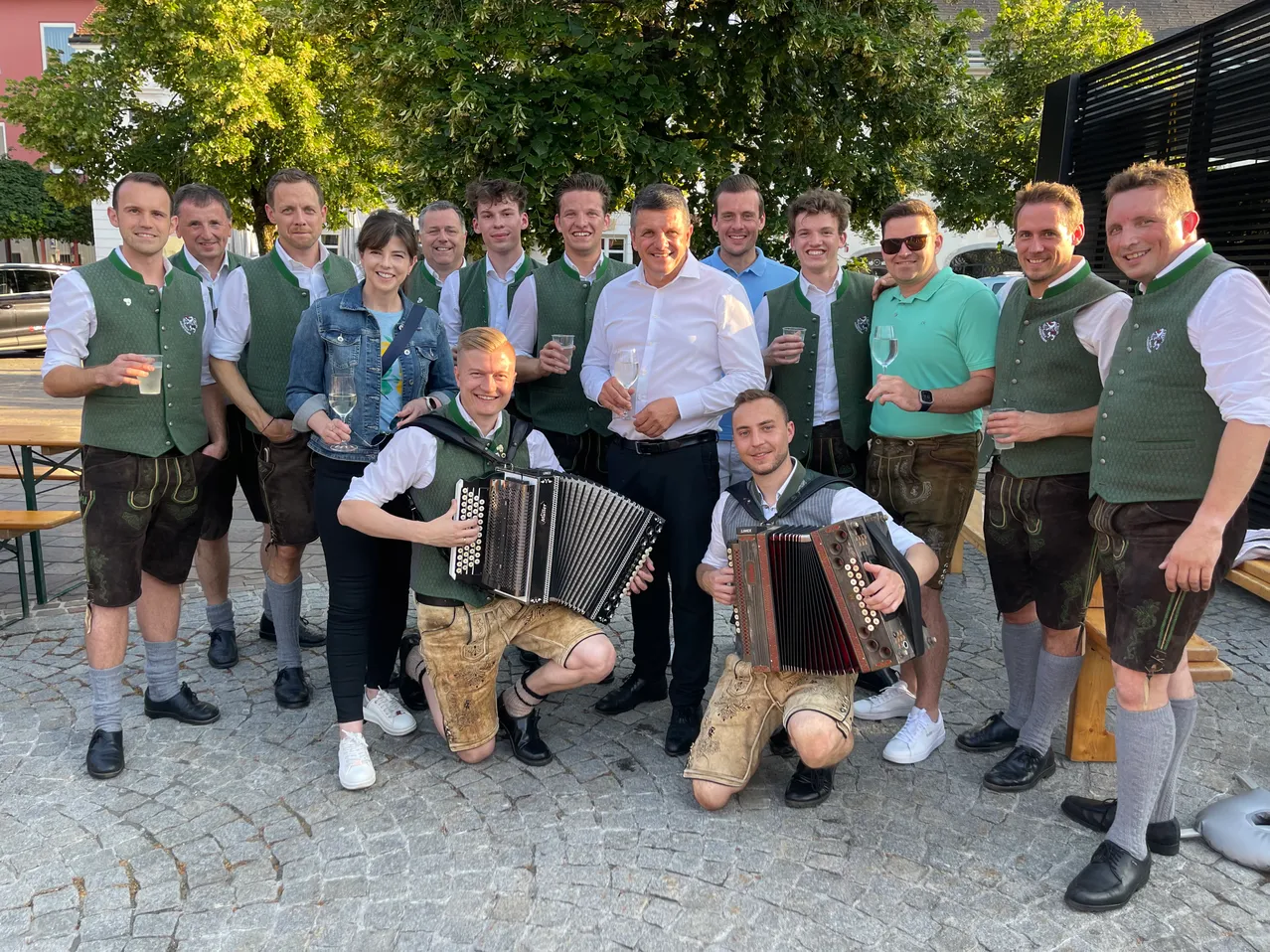 Eine Gruppe von Menschen in traditioneller Kleidung steht lächelnd im Kreis, zwei Männer spielen Akkordeons. Im Hintergrund sind Bäume und ein Gebäude zu sehen.