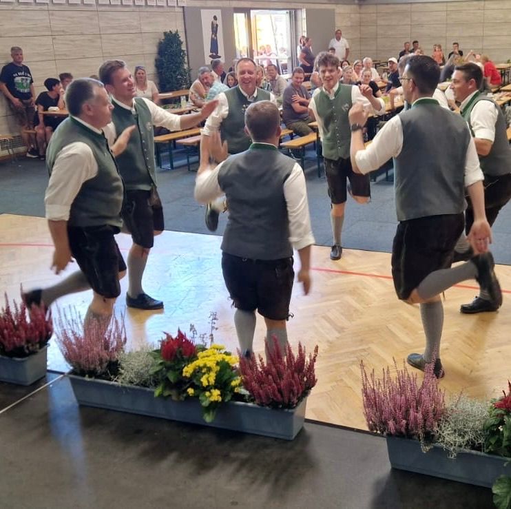 Eine Gruppe von Männern in traditioneller deutscher Kleidung tanzt auf einer Bühne mit Holzboden und Blumen. Das Publikum beobachtet sie im Hintergrund.