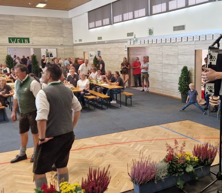 Eine Indoor-Veranstaltung mit mehreren Personen an Tischen, andere stehen. Einige Leute tragen traditionelle Kleidung. Topfpflanzen stehen auf dem Boden und an den Wänden.