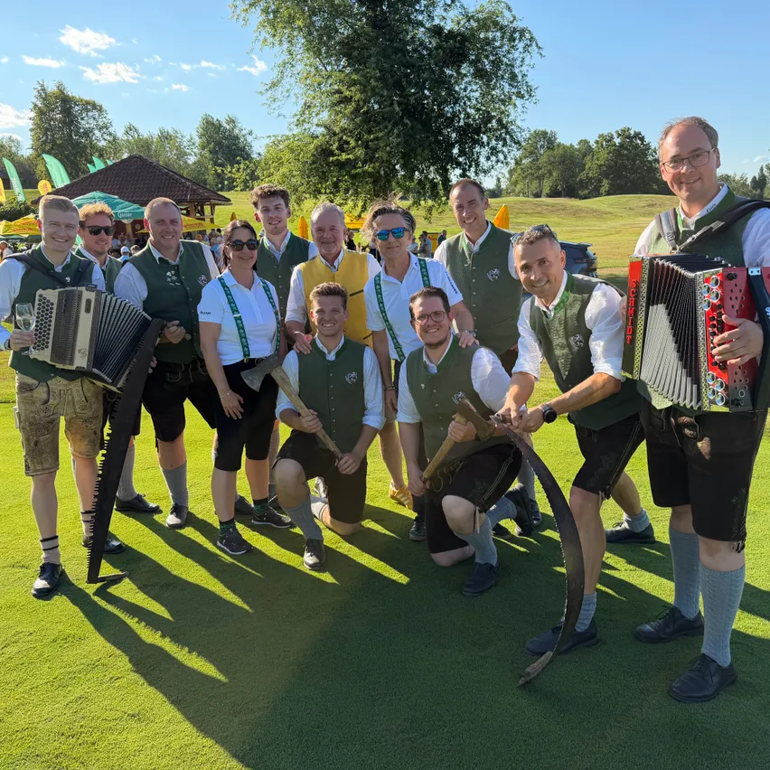 Eine Gruppe von Musikern in traditioneller Kleidung, einschließlich grüner Westen und Shorts, posiert mit Musikinstrumenten wie Akkordeons und Sägen auf einem grünen Feld.