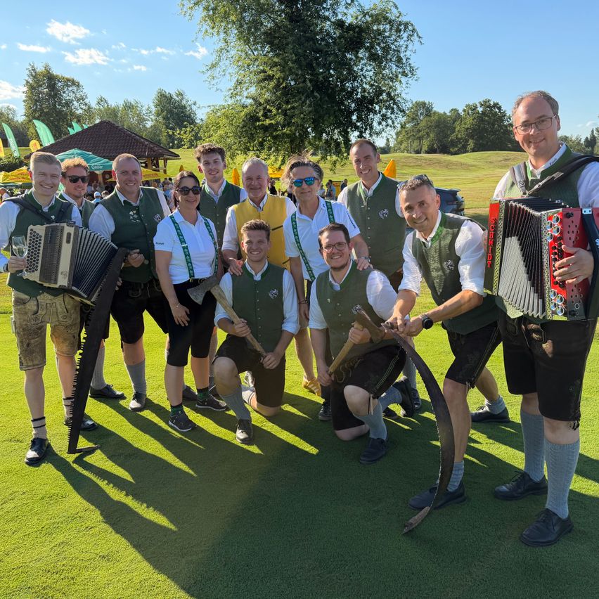 Eine Gruppe von Musikern in traditioneller Kleidung, einschließlich grüner Westen und Shorts, posiert mit Musikinstrumenten wie Akkordeons und Sägen auf einem grünen Feld.