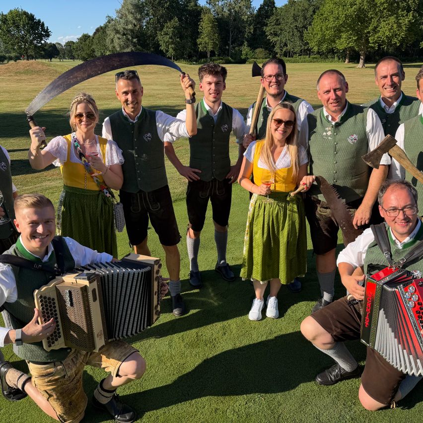 Eine Gruppe von Menschen in traditioneller Kleidung posiert mit Musikinstrumenten und Äxten auf einer Wiese.