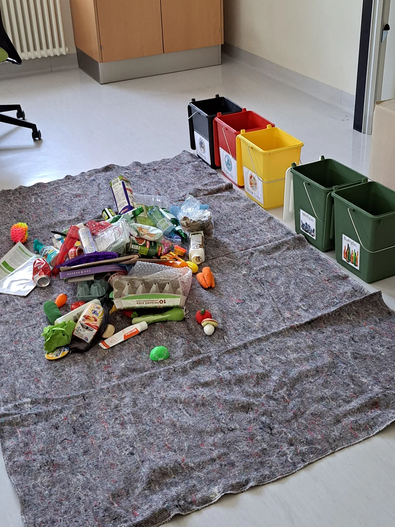 Ein grauer Bodenteppich zeigt eine Sammlung verschiedener Wertstoffe. Verschiedene Gegenstände wie Plastik, Papier und Lebensmittelabfälle sind ordentlich angeordnet. Vier farbige Behälter sind für verschiedene Arten von Abfällen gekennzeichnet.