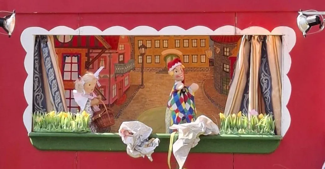 Eine Puppenszene zeigt zwei Figuren in einer bunten Stadtlandschaft, eine in einem Clownkostüm. Die Szene umfasst Gebäude, eine Straßenlaterne und Blumen.