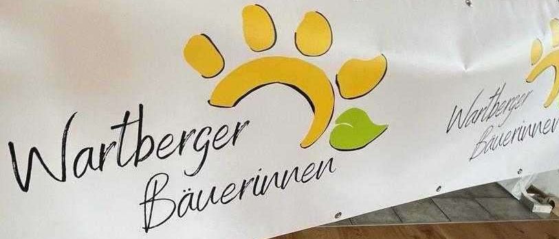 Ein weißer Banner mit gelben Pfotenabdrücken und grünen Blättern trägt den Text 'Wartberger Bauerinnen'. Dahinter ist ein Raum mit Holzböden, einem Glasschrank und einem Fenster mit Vorhängen zu sehen.