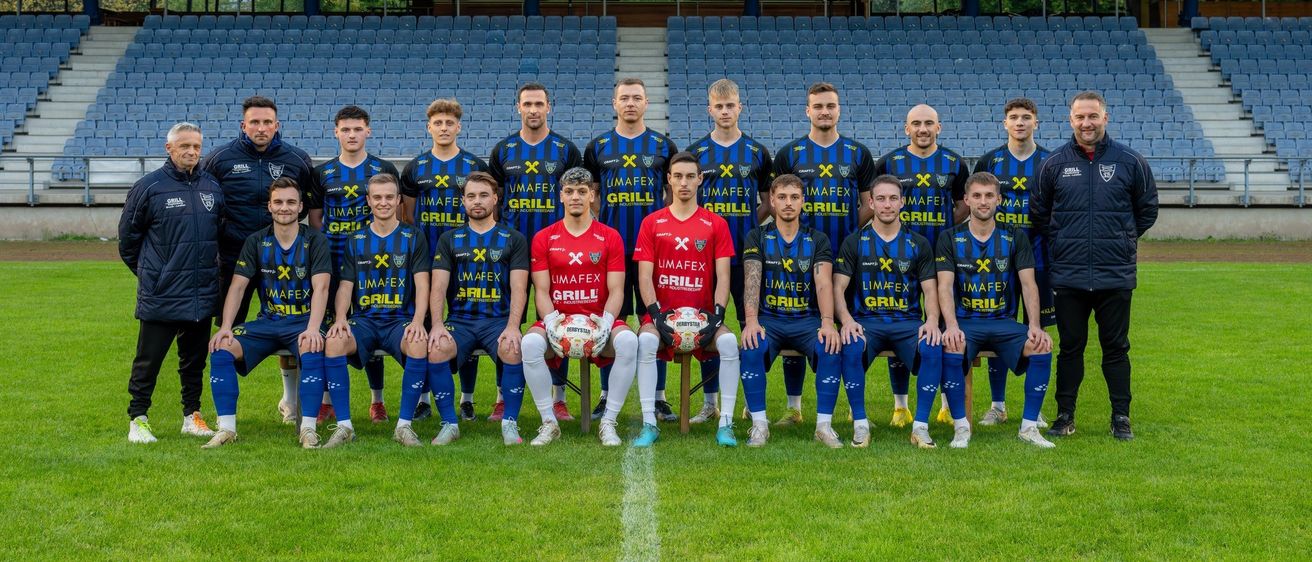 Eine Fußballmannschaft posiert für ein Foto auf einem Feld, in blauen und gelben Uniformen. Die Spieler lächeln und halten Fußballbälle. Das Stadion hat leere blaue Sitze im Hintergrund.