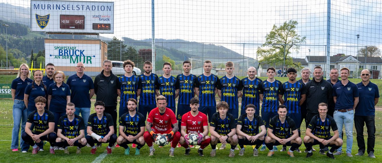 Eine Fußballmannschaft posiert für ein Gruppenfoto auf einem Feld mit Netz. Sie tragen blaue Uniformen mit roten Säumen. Die Anzeigetafel zeigt Heim 0, Gast 1. Bäume und Berge sind im Hintergrund.