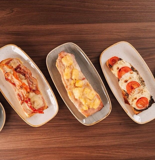 Drei rechteckige Teller auf einer Holzoberfläche zeigen verschiedene Arten von Sandwiches. Ein Sandwich hat Speck und Käse, ein anderes eine gelbe Zutat und das letzte ein gebratenes Ei und Tomatenscheiben.