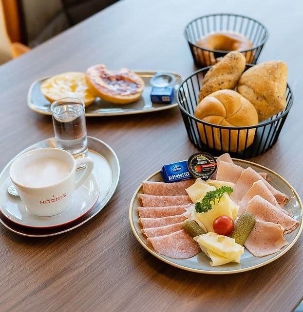 Ein Frühstück auf einem Holztisch umfasst eine Tasse Kaffee, Brot, Käse, Gurken, geschnittenes Fleisch. Andere Gegenstände sind ein Glas Wasser und ein Tablett mit einem Bagel.