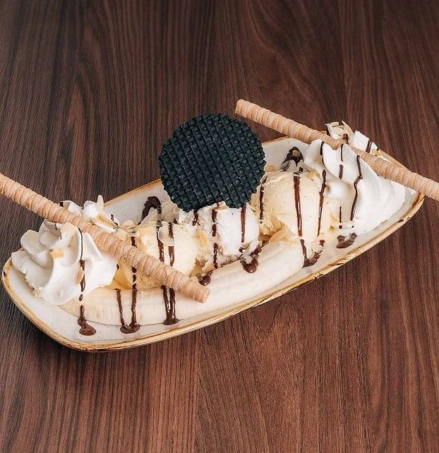Ein ovaler Teller hat einen Bananensplit, der mit Eiscreme, Schlagsahne und Schokoladensirup garniert ist und mit einem Waffelkegel und zwei Spießen verziert ist.