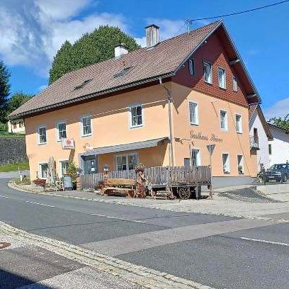 Eine Straße mit einem Gebäude an der Seite. Einige Bänke und ein Wagen sind draußen. Ein Auto ist vor dem Gebäude geparkt. Bäume und Büsche sind in der Umgebung.