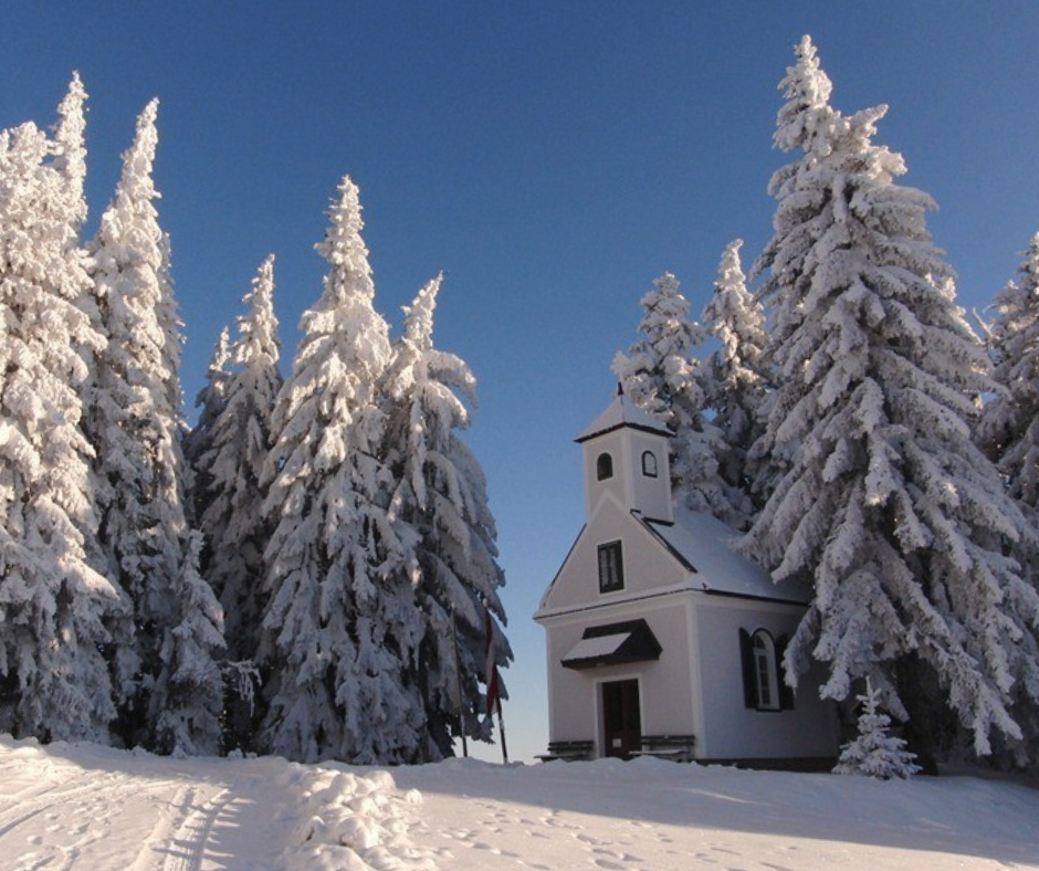 Eine kleine weiße Kirche, bedeckt mit Schnee, steht zwischen hohen Tannen in einer verschneiten Landschaft.