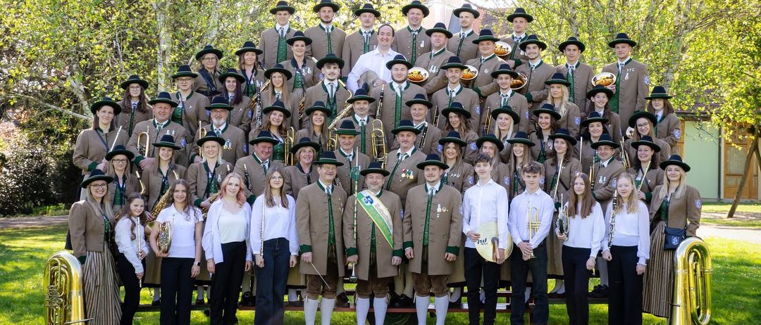 Eine große Gruppe von Musikern in traditioneller Kleidung posiert für ein Foto auf einem Rasenfeld mit Bäumen im Hintergrund. Sie halten verschiedene Instrumente wie Trompeten, Posaunen und Klarinetten.