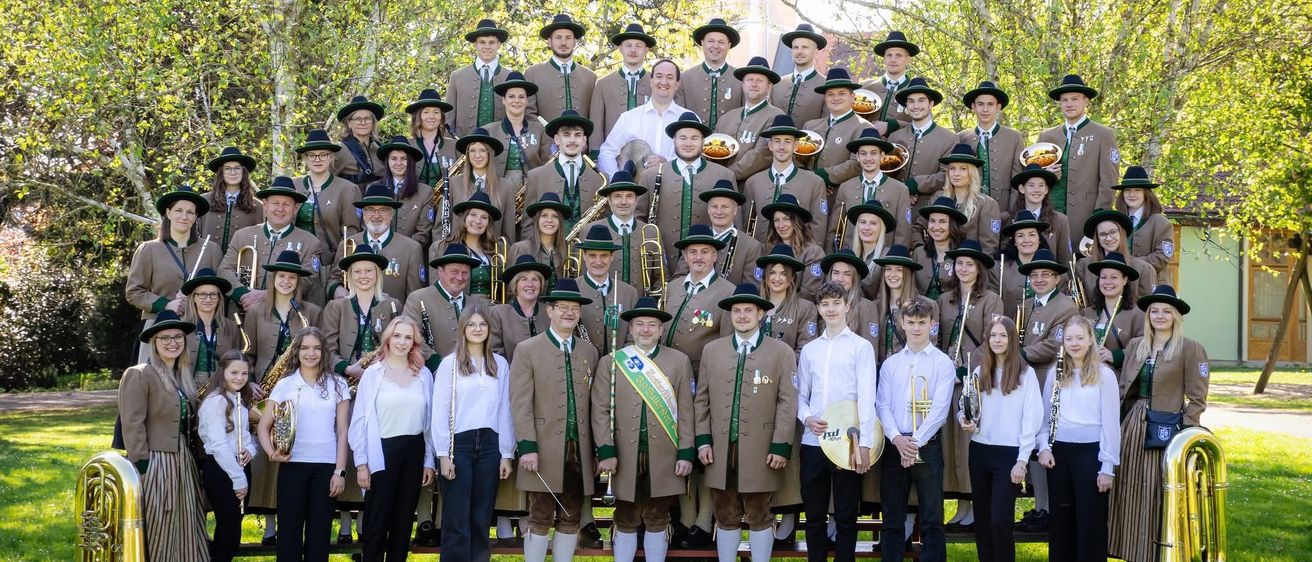 Eine große Gruppe von Musikern, gekleidet in übereinstimmenden Uniformen, steht vor einer Reihe von Bäumen und einem Grasfeld. Sie halten verschiedene Musikinstrumente in den Händen, darunter Trompeten, Posaunen und Flöten. Einige tragen Hüte und Schuhe, während andere barfuß sind. Die Gruppe scheint für ein Foto zu posieren.