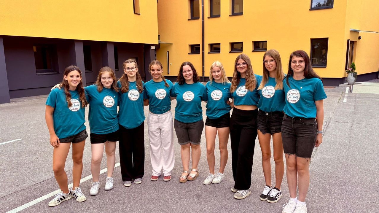 Eine Gruppe junger Frauen in passenden türkisen Shirts mit einem Logo steht vor einem gelben Gebäude. Sie lächeln und scheinen für ein Foto zu posieren.