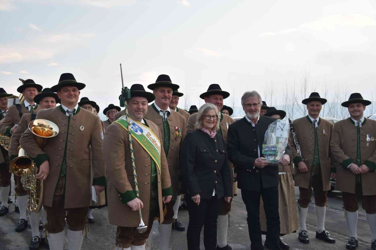 Eine Gruppe von Menschen in traditioneller bayerischer Kleidung, einschließlich Hüten, Medaillen und Schärpen, steht zusammen im Freien, wobei ein Mann einen Pokal hält.