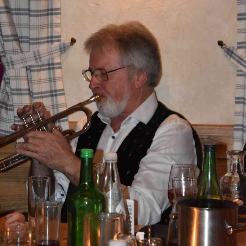 Ein älterer Mann mit Brille sitzt an einem Tisch und spielt Trompete. Flaschen und Gläser stehen auf dem Tisch, vor ihm ein Krug und eine Schüssel.