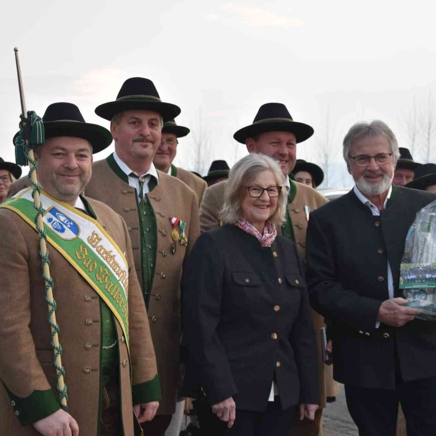 Eine Gruppe von Menschen ist in traditioneller Festkleidung gekleidet. Ein Mann vorn trägt ein grünes Band und hält einen Stab. Der Mann neben ihm trägt einen braunen Mantel mit Medaillen. Die Frau in der Mitte trägt ein Halstuch und eine Brille. Der Mann rechts hält eine Plastiktüte.