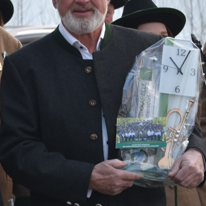 Ein Mann in einem Anzug hält ein verpacktes Geschenk mit einer Uhr darin, die 1:51 anzeigt. Hinter ihm tragen andere Hüte und Jacken.