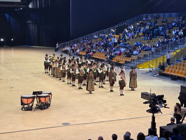 Eine Blaskapelle in traditioneller Kleidung tritt auf einer Bühne in einer überdachten Arena auf, mit einem Publikum in den Rängen.