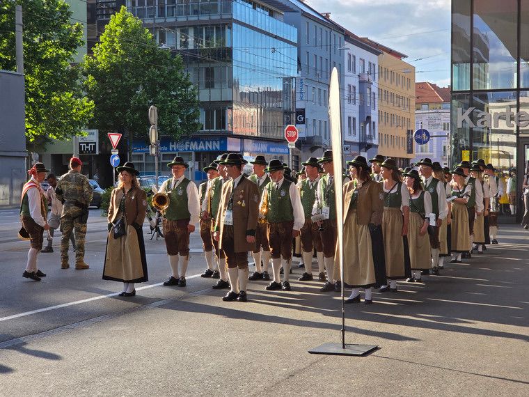 Eine Gruppe von Menschen in traditioneller bayerischer Kleidung, einschließlich grüner Westen und Hüten, marschiert in einer Parade auf einer Stadtstraße. Hinter ihnen geht ein Soldat in Tarnkleidung. Im Hintergrund sind Gebäude, Bäume und Straßenschilder zu sehen.