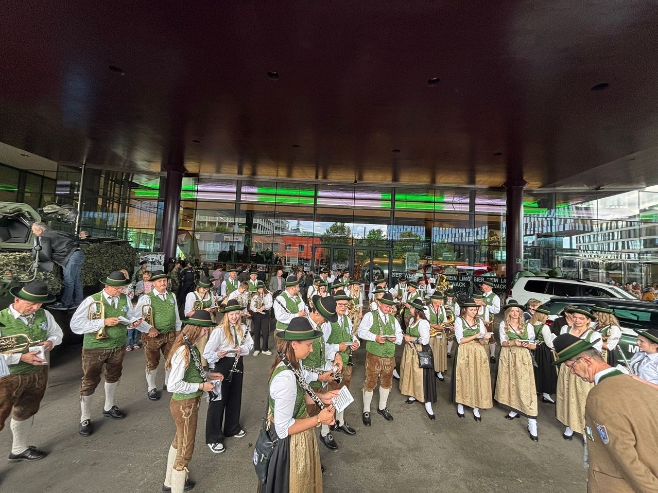 Eine Gruppe von Musikern, die in traditioneller deutscher Kleidung, einschließlich grüner Westen und Hüten, gekleidet sind, versammelt sich in einem modernen Gebäude mit großen Glaswänden und grünen Lichtern. Sie halten Musikinstrumente und scheinen sich auf eine Aufführung vorzubereiten.