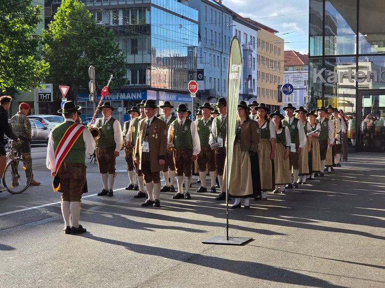 Eine Gruppe von Menschen in traditioneller bayerischer Kleidung marschiert bei einer Parade auf einer Stadtstraße. Sie werden von einem Soldaten und einem Radfahrer begleitet. Die Gebäude im Hintergrund sind modern und haben Glasfassaden.