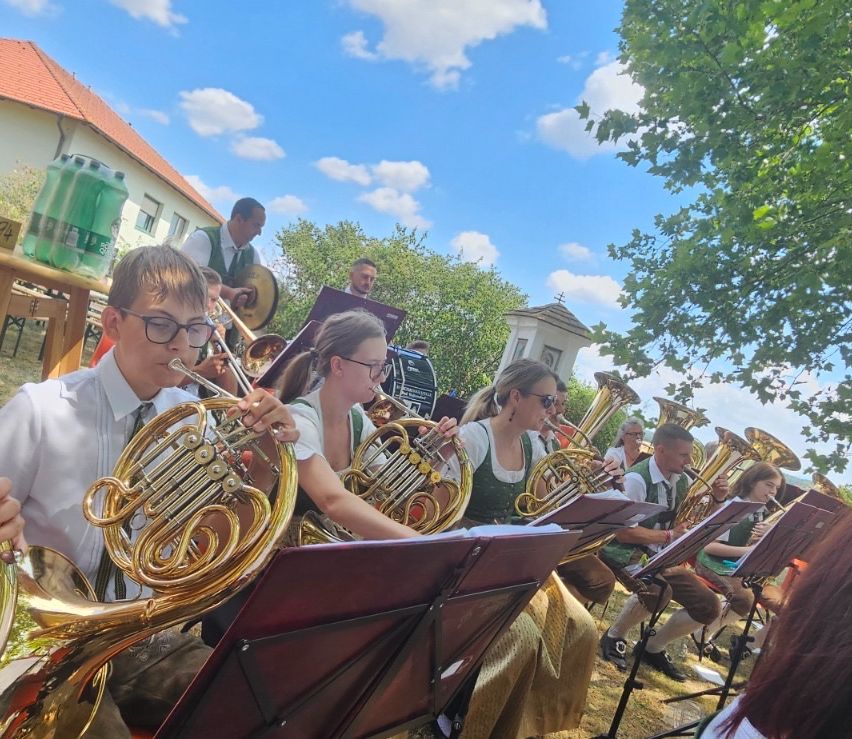 Eine Gruppe von Musikern, darunter ein Junge und Mädchen, spielen Blechblasinstrumente im Freien. Sie sitzen auf dem Gras mit Notenständern vor ihnen. Dahinter steht ein Gebäude mit einem roten Dach und einem Baum.