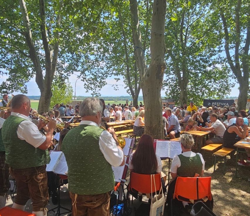 Eine Gruppe von Musikern in traditioneller bayerischer Kleidung spielt auf einer Bühne in einem Freiluftpark, während eine Menge Menschen auf Bänken und Tischen sitzt und die Musik genießt.