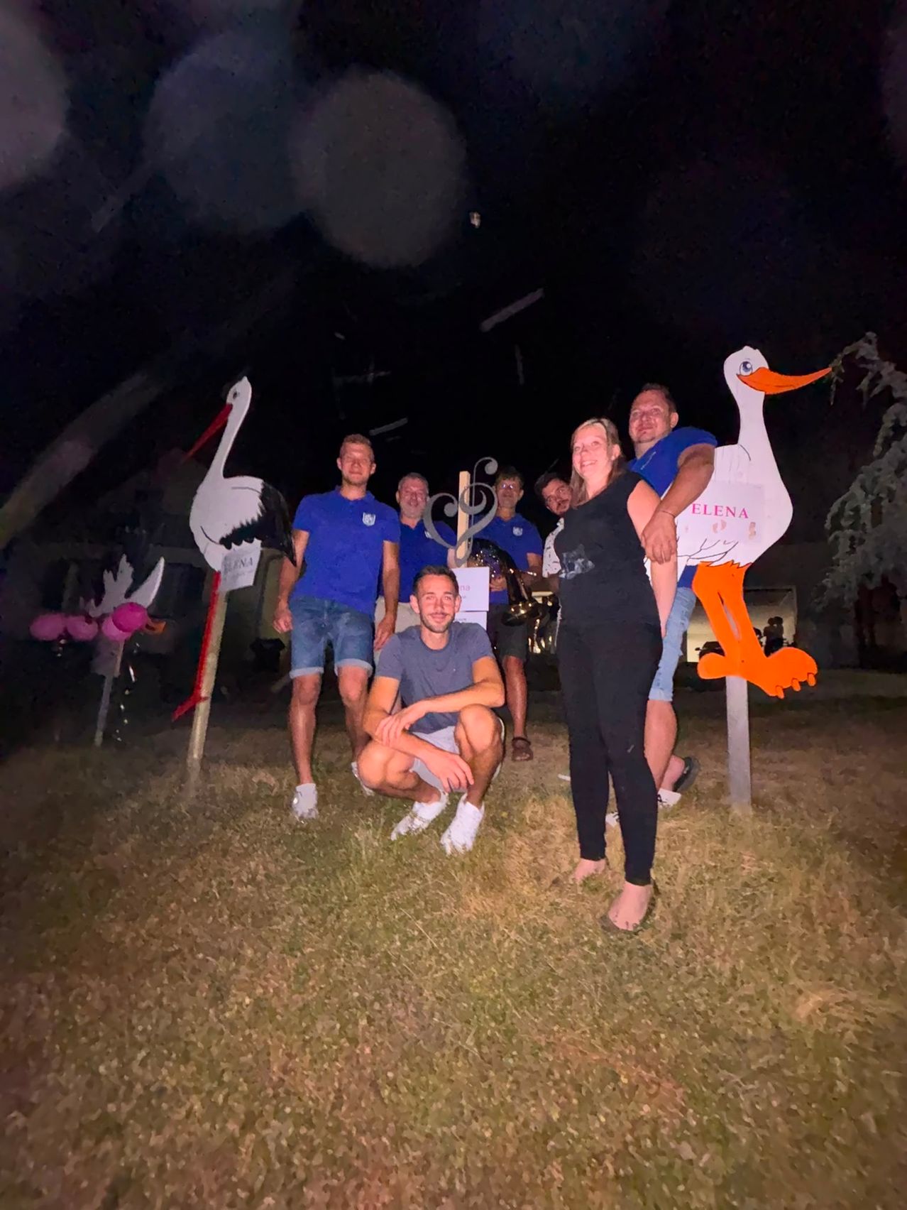 A group of people are posing for a photo at night in front of large stork cutouts. Some are standing, while others are sitting on the ground.