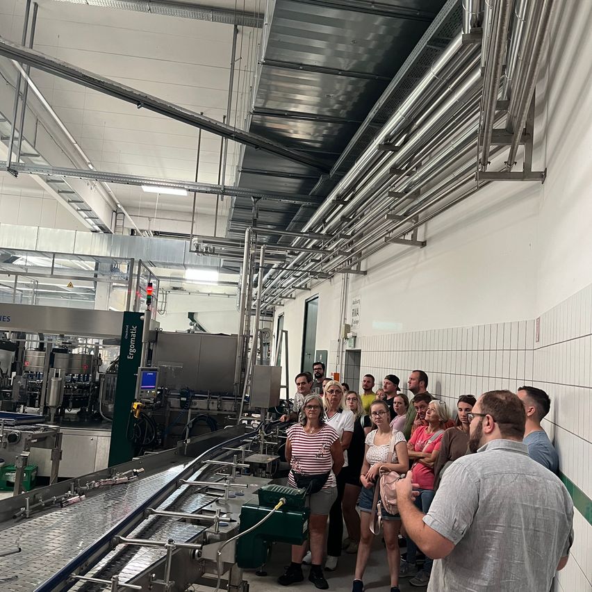 Eine Gruppe von Menschen steht in einer Fabrik und hört einem Mann zu, der spricht. Die Fabrik hat ein Förderband und Maschinen.