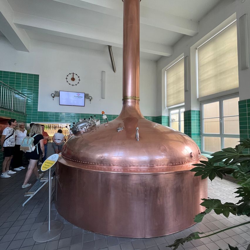 Ein großer Kupfer-Braukasten in einer Brauerei, um den herum Menschen stehen. Der Kessel ist von grünen Fliesen umgeben und hat eine Uhr an der Wand.