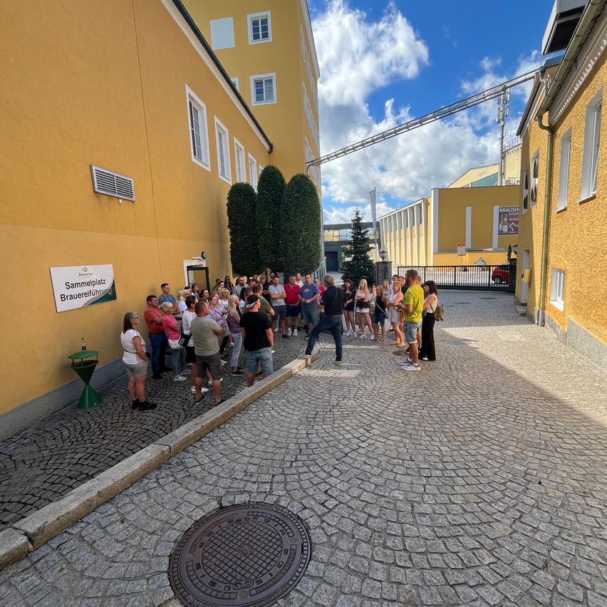 Eine Gruppe von Menschen versammelt sich vor einem gelben Gebäude mit einem Schild, das Sammelplatz Brauereiführung steht. Sie stehen auf einer Kopfsteinpflasterstraße unter einem teilweise bewölkten Himmel.