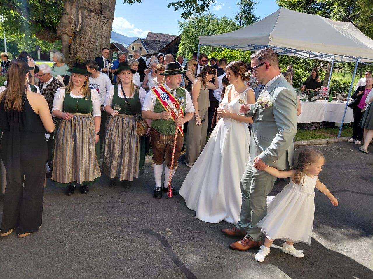 Eine Hochzeitsfeier in traditioneller Kleidung steht auf einem Freigelände mit einem Zelt. Die Braut und der Bräutigam halten Händchen mit einem Kind. In der Nähe beobachten Menschen in Anzügen und Kleidern. Bäume und Häuser sind im Hintergrund.